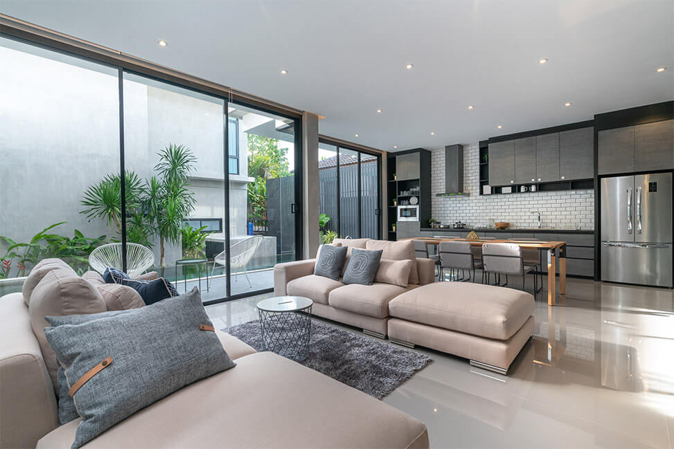 Contemporary open-plan, organized and cleaned living room with large windows, neutral-toned sofas, modern kitchen in the background, and indoor greenery