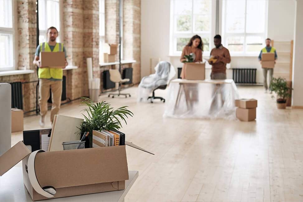 Moving day scene with people carrying boxes in a spacious, sunlit room, featuring large windows and a sofa in the foreground.