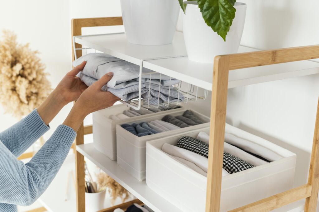 An organized storage shelf in a light and airy setting. A person's hands are seen neatly folding and placing a piece of clothing on one of the shelves. Below, there are various clothes compartmentalized into plastic storage bins. The color scheme is neutral with elements of white and light wood. The decor is complemented by plants and dried flowers, giving the space a fresh and serene atmosphere.