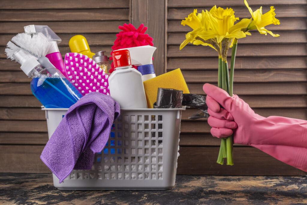 Spring cleaning supplies in a basket with vibrant yellow flowers held by hand in pink gloves against a wooden background.