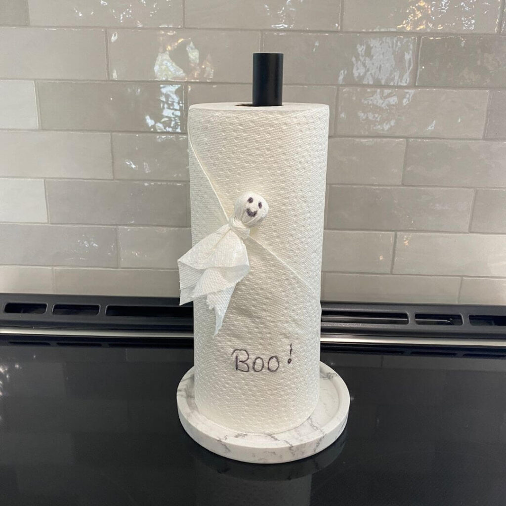 That's a fun and creative Halloween-themed decoration! It looks like a paper towel roll adorned with a small ghost made out of a white tissue or cloth, complete with a drawn face. The ghost is tied to the roll with a white ribbon, and there's a handwritten "Boo!" on the paper towel. It's placed on a countertop with a gray tiled backsplash in the background. Such a cute and simple idea to bring some festive spirit to the kitchen!
