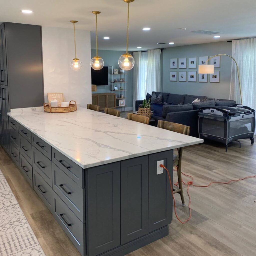 A spacious open-concept living area with a prominent kitchen island in the foreground. The island has a white marbled countertop, and its base is painted dark gray with multiple drawers and cabinet space. Above the island hang three large globe pendant lights with gold fixtures. To the left, tall dark gray cabinets match the color of the island. To the right, the living area is furnished with a deep blue sectional sofa, behind which is a wall adorned with a grid of framed pictures. In the corner, there's a modern arched floor lamp with a gold finish, next to a baby playpen. The room is well-lit and has light wooden flooring with a beige patterned rug visible in the foreground.