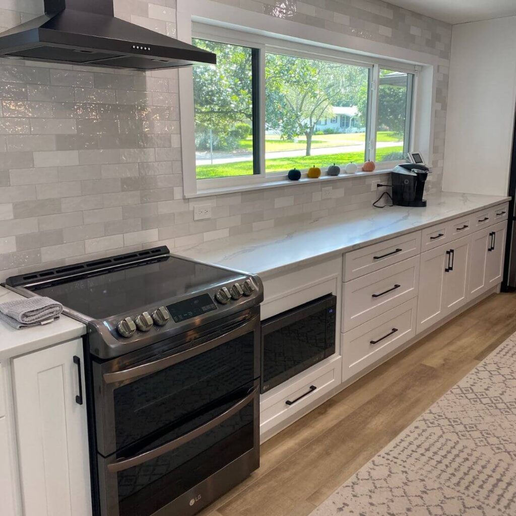 A close-up view of a section of a modern kitchen. On the left, there's a stainless-steel range oven with a black cooktop, digital controls, and silver knobs. Above it is a sleek black exhaust hood. The backsplash is made of white subway tiles. On the countertop, which has a marbled design, a series of colorful fruit sits in front of a large window that offers a view of a lush green lawn and trees outside. On the right, there's a black coffee machine, and below are white cabinets with black handles. At the bottom right corner, a beige patterned runner is partially visible on the floor.