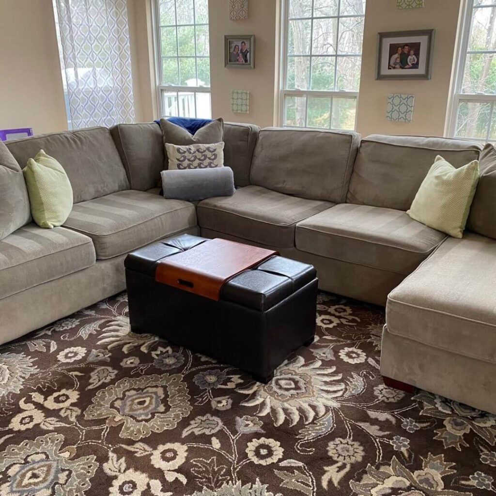 Cozy living room interior with a gray sectional sofa, patterned area rug, leather ottoman, and decorative pillows, surrounded by framed photos on the wall and large windows.