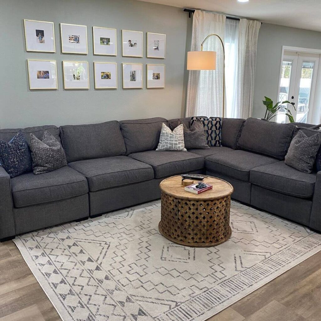 A cozy living room featuring a large gray sectional sofa adorned with various patterned pillows. Above the sofa, a set of twelve framed photographs are symmetrically displayed on the wall. In front of the sofa, there's a round, wooden carved coffee table with a few magazines on top. The room has a geometric-patterned gray and white rug. To the right, a modern floor lamp with a large white shade arches over the sofa, and there's a glimpse of a window with sheer white curtains.