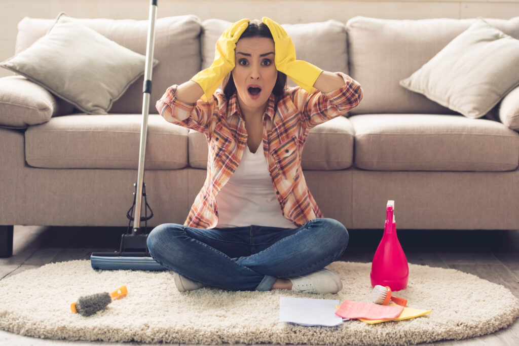 Woman wearing yellow gloves expressing surprise while sitting on the floor, with cleaning supplies scattered around and a mop beside her, in front of a couch.