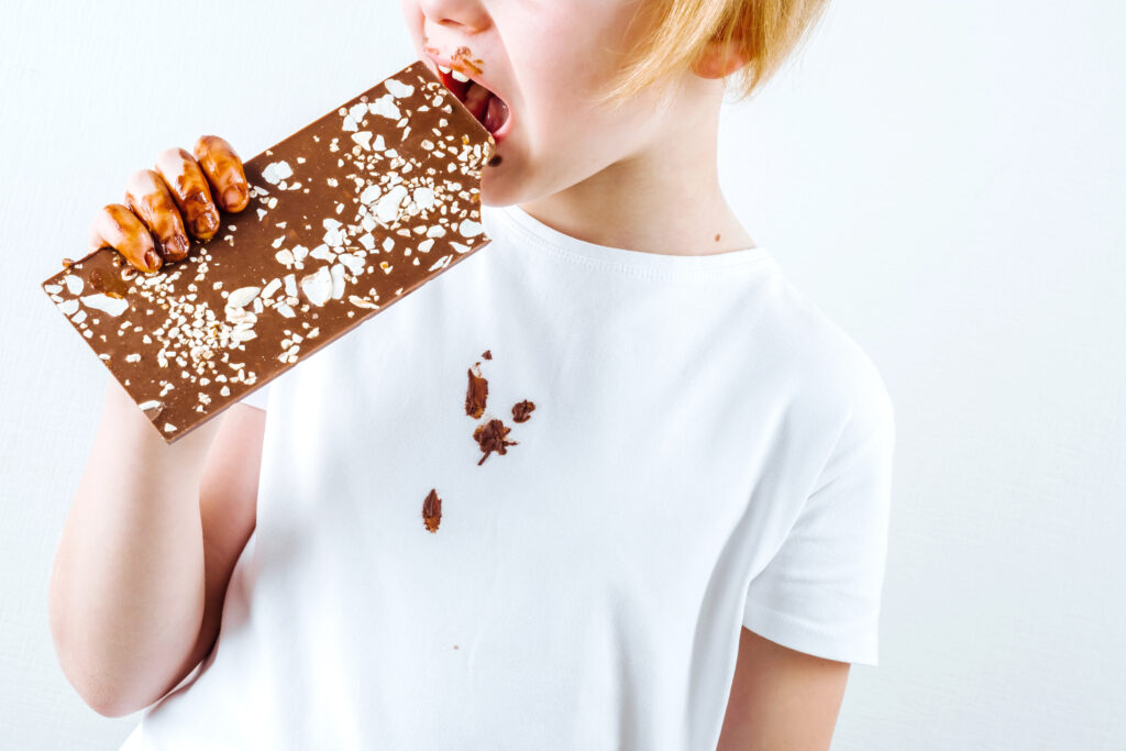 Child in a white t-shirt enjoying a large chocolate bar with hazelnuts, with chocolate smudges on face and shirt, representing the joy of sweet treats.