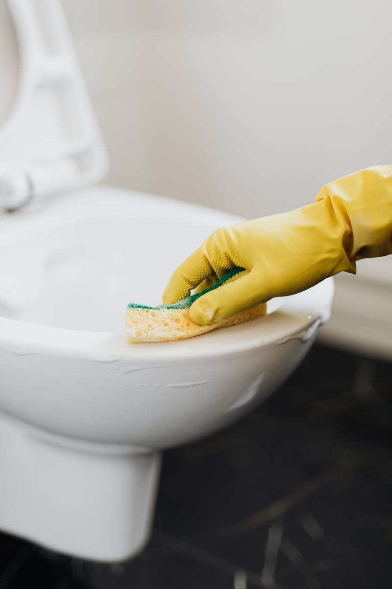 Crop anonymous person wearing yellow cleaning glove removing stains on toilet bowl with sponge in washroom