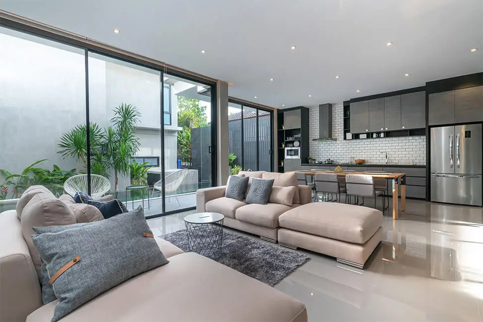 Contemporary open-plan, organized and cleaned living room with large windows, neutral-toned sofas, modern kitchen in the background, and indoor greenery