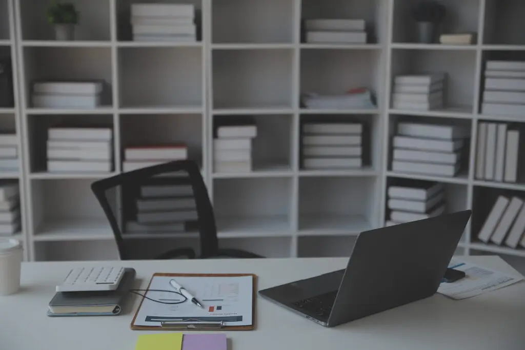 A modern office workspace with a white bookshelf filled with books, a desk containing a laptop, calculator, notepad, and other office supplies, and a black office chair. The color scheme is predominantly neutral, and the atmosphere is clean and organized.