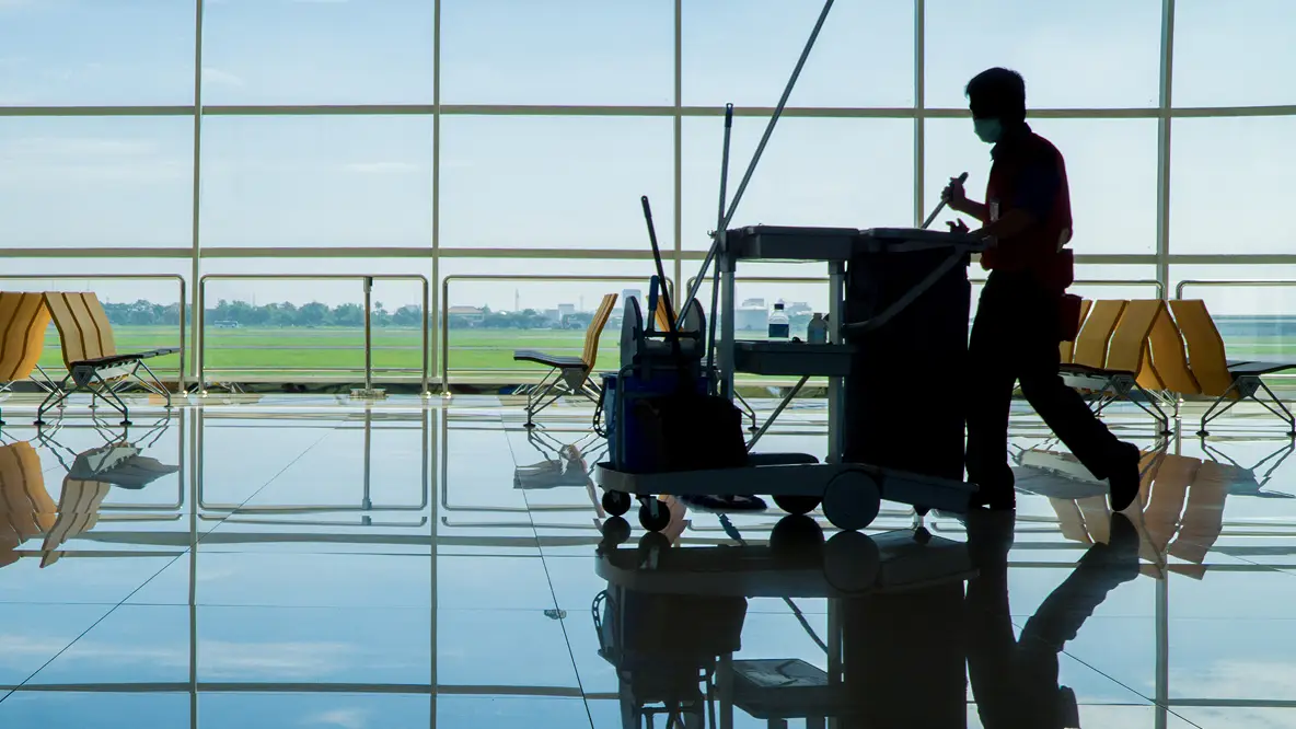 A janitor in silhouette pushes a cleaning cart in an empty airport terminal with rows of yellow seats and large windows overlooking a runway.