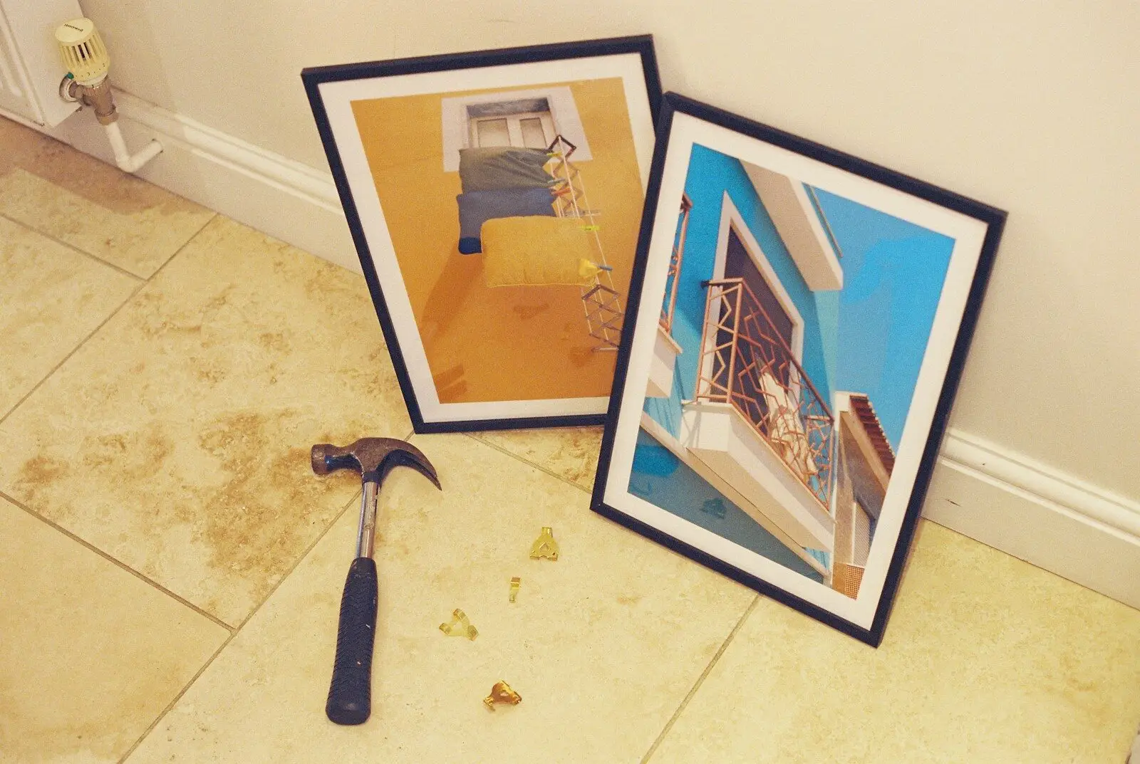 A hammer and brass picture hooks on a tiled floor near two framed prints